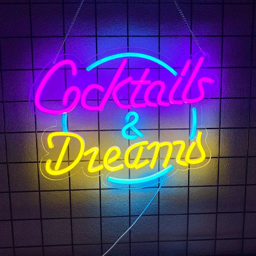 A neon sign reading 'Cocktails & Dreams' in glowing pink, yellow, and cyan blue lettering, mounted against a dark tiled wall.