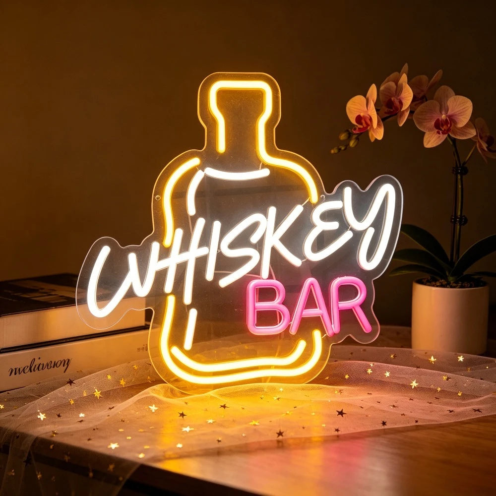 A glowing decorative sign shaped like a whiskey bottle with the words “WHISKEY BAR” in white and pink neon-style lettering, displayed on a tabletop in a warmly lit indoor setting with a potted orchid in the background.